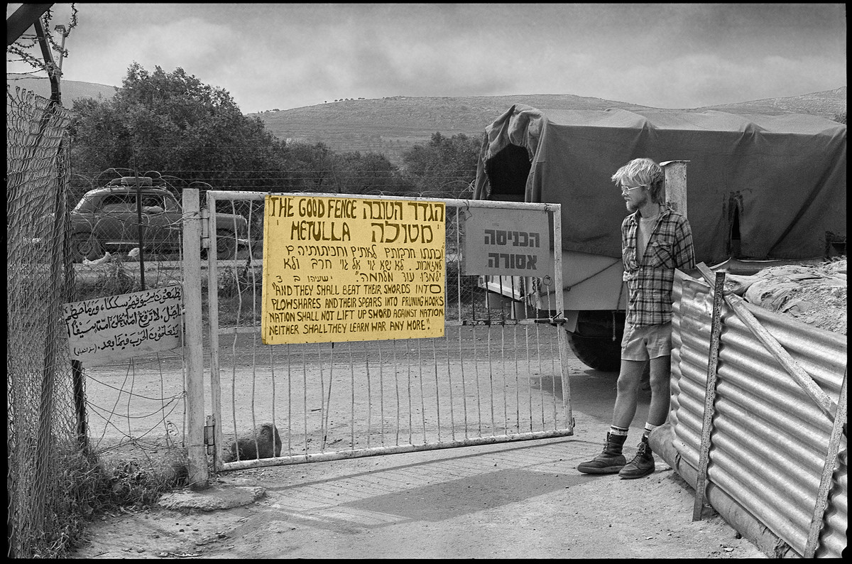 Young man stands in front of gate with sign reading The Good Fence