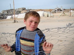 Boy leans against fence with destroyed homes in background