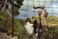 Man with machete makes hole in barbed wire fence as couple dressed as bride and groom stand in background in natural landscape