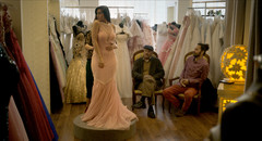 Woman wearing beaded gown stands on pedestal as two men sitting on chairs look on in bridal dress shop