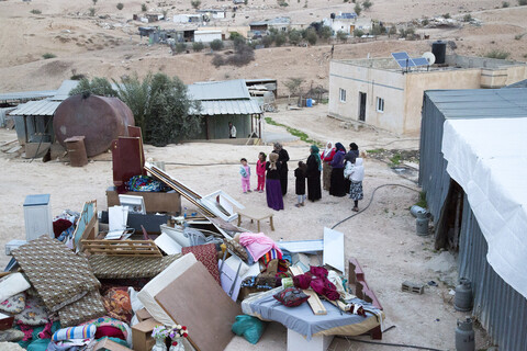 Women and children stand near home with possessions piled up outside