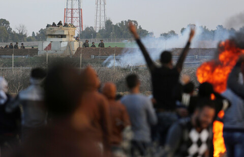 Israeli soldiers in watchtower and along fence look on in background as Palestinian youths with backs to their camera are seen in foreground