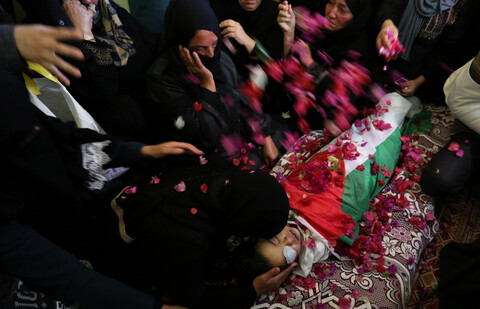 Women lean over and throw flower petals on body of boy shrouded in Palestinian flag