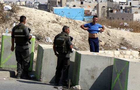Two soldiers behind concrete blocks look at a young man who has lifted his t-shirt to show his stomach