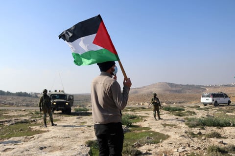 Man with back turned to camera holds Palestine flag while facing soldiers