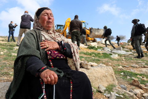 Woman sits on a rock with her hand on her chest as bulldozer operates in the background 