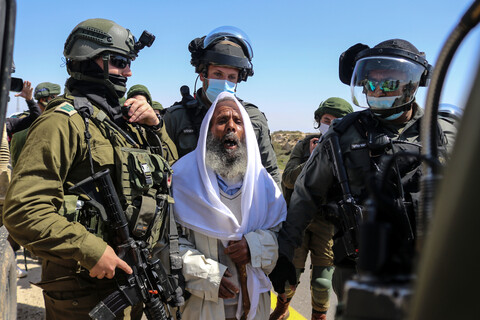 An older man clutches a white shawl worn over his head while soldiers stand around him