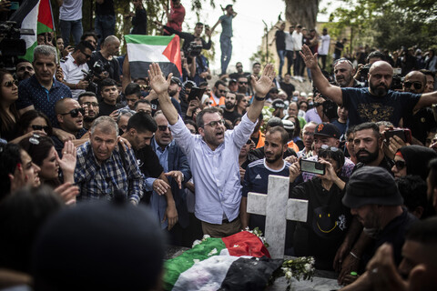 Man holds his hands above his head while standing in crowd around grave with Palestine flag on it