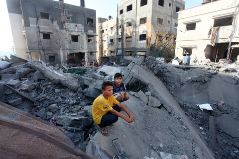 Two boys sit on ruins of fallen building while surrounded by bombed-out midrise buildings