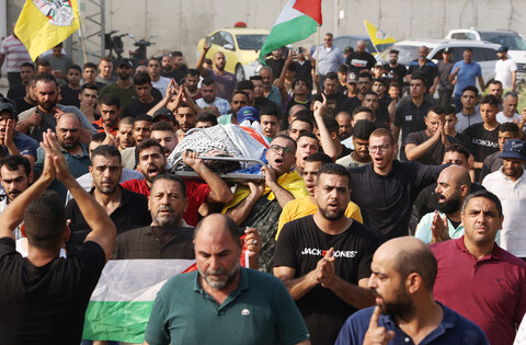 Body shrouded in kuffiyeh and Palestine flag is carried on stretcher by crowd of men, some carrying Palestine and Fatah flags