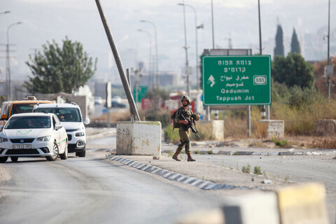 Soldier carrying rifle walks on median with Palestinian-plated cars in one lane and an empty lane on the other side