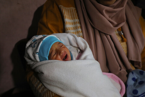 A baby wearing a blue cap is swaddled in a white blanket