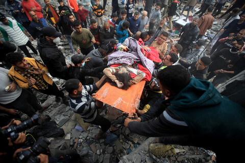 A child partly covered by a blanket is carried on a stretcher by several people amid a large crowd