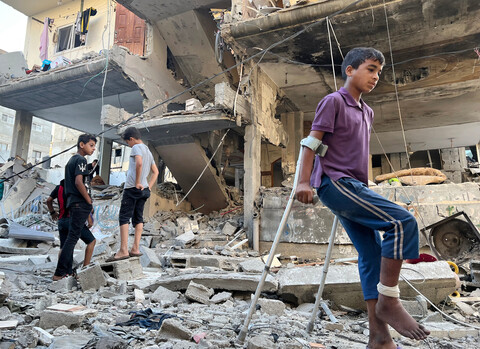 A boy with a bandaged leg uses crutches while walking across rubble in front off bombed-out building