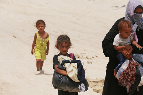 A woman holds a baby while two other children walk beside or behind her 