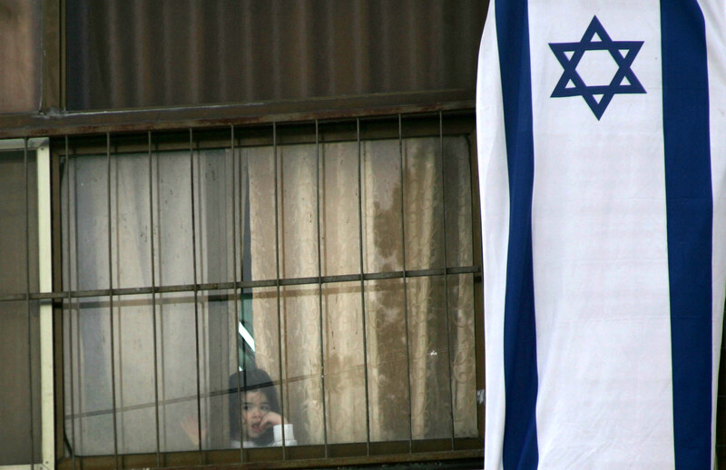 Small girl peers out of window covered with giant Israeli flag