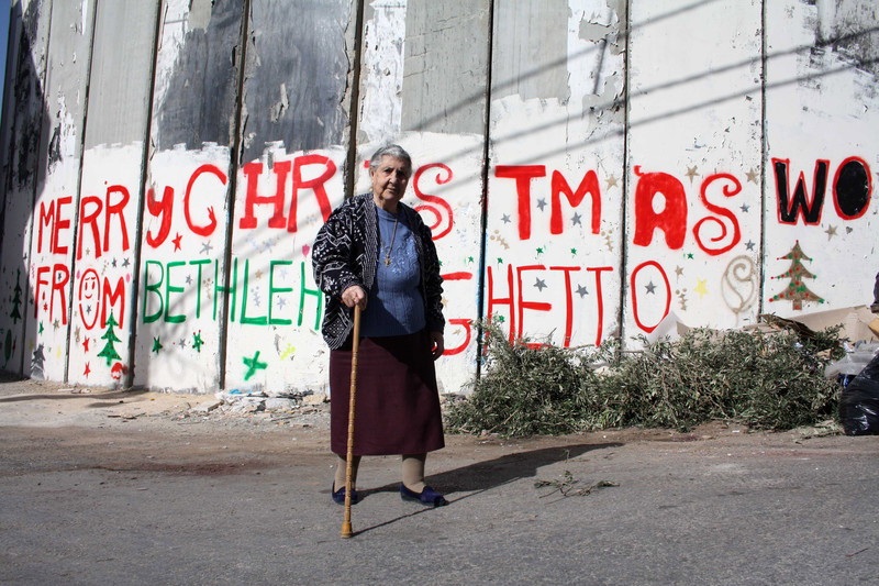 Woman walks in front of wall reading &quot;merry Christmas from Bethlehem Ghetto&quot;