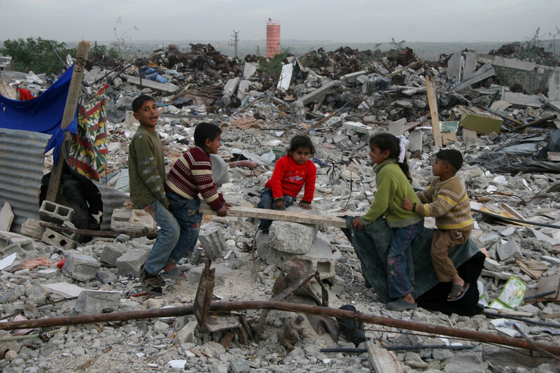 Children play on improvised see-saw amidst rubble of destroyed buildings