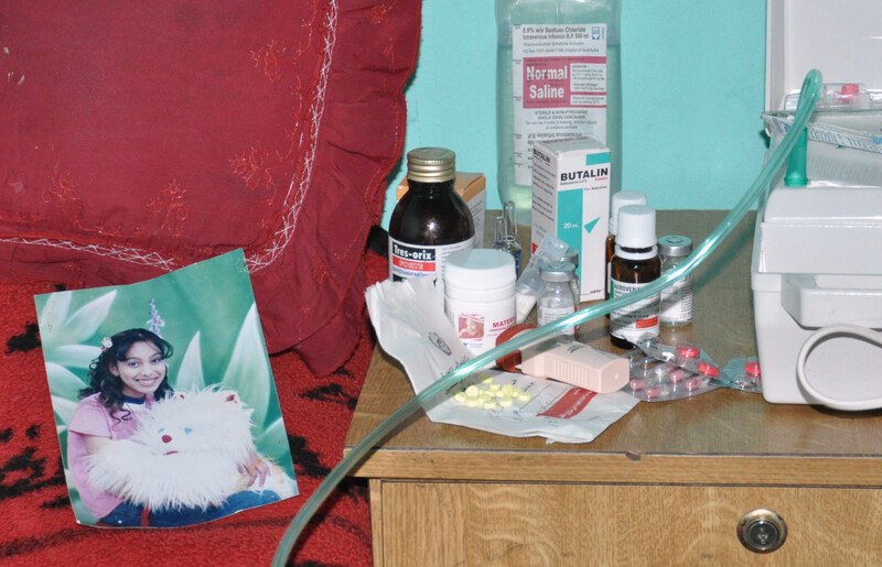 A portrait photo of Haneen lays on her bed while medicines sit on table next to bed