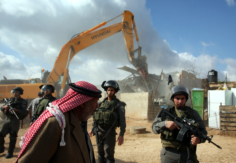 Bedouin man walks in front of Israeli soldiers guarding Hyundai equipment destroying structures