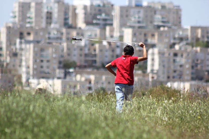 Boy with slingshot in field with settlement mid-rises in background