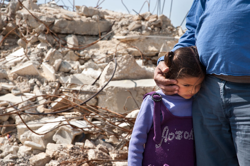 Close-up of man cradling head of girl, with rubble of destroyed home in background