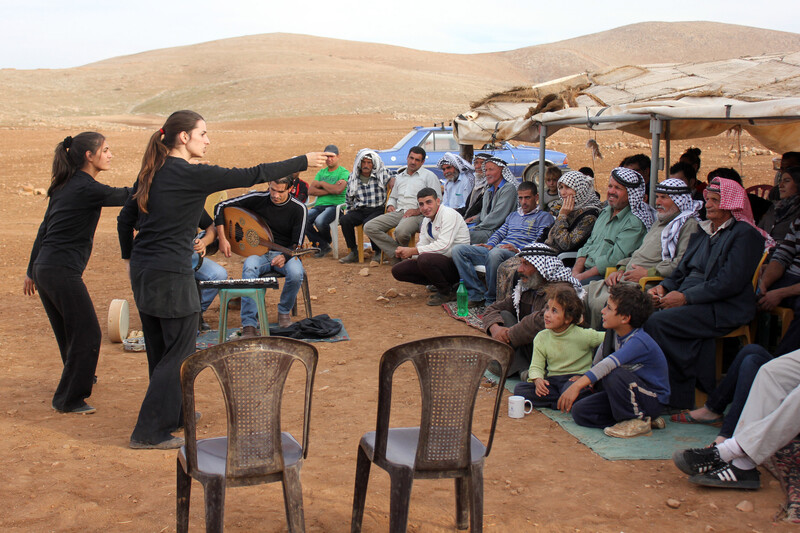 Women in black gesture towards audience in outdoor setting