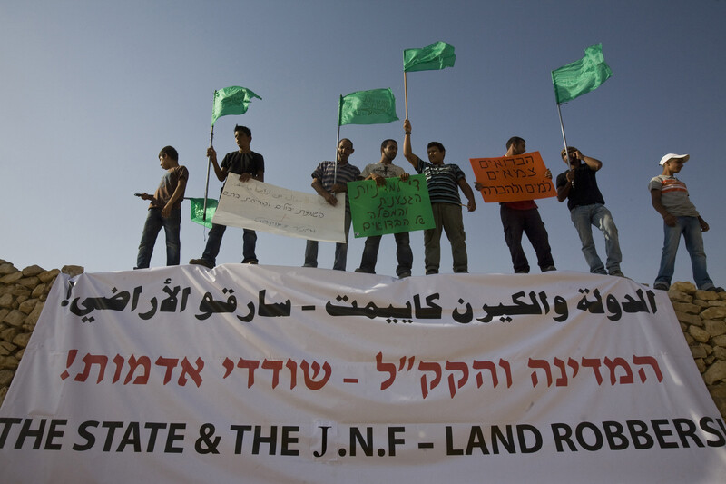 Men carrying signs and flags stand above banner reading "State & the JNF - Land Robbers" in Arabic, Hebrew and English