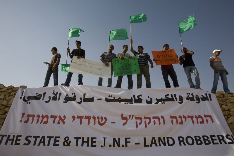 Men carrying signs and flags stand above banner reading &quot;State &amp; the JNF - Land Robbers&quot; in Arabic, Hebrew and English