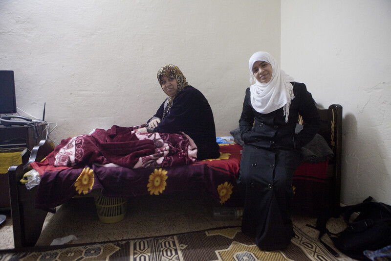 Older woman sitting on bed and and younger woman standing in front of bed smile at camera