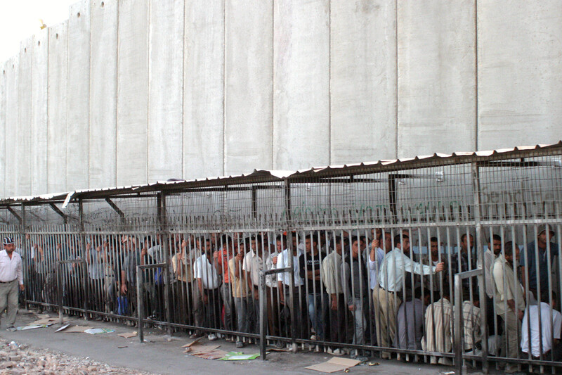 Palestinian men queue at checkpoint next to very tall concrete wall