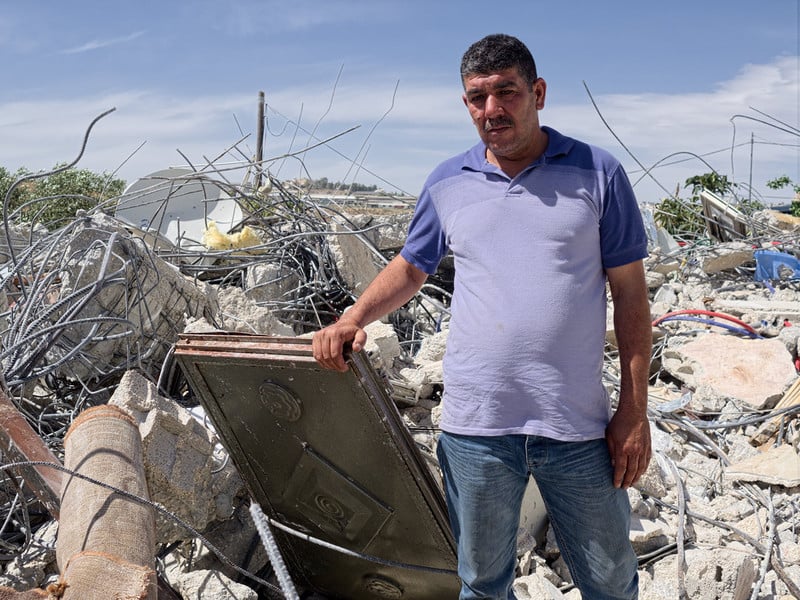 Man holds piece of the rubble of his home, seen in background