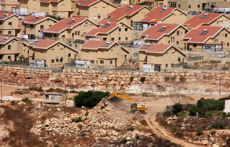 An earth-moving machine works in the foreground with Israeli settlement homes in background