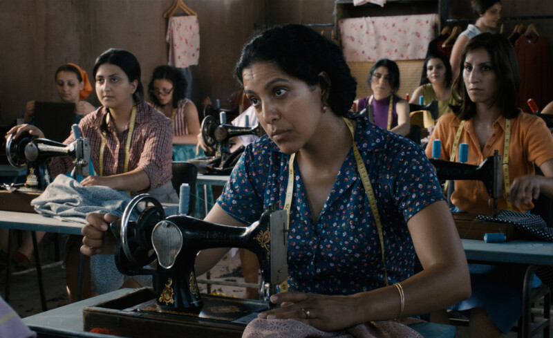 Close up of woman sitting behind sewing machine in room full of seamstresses