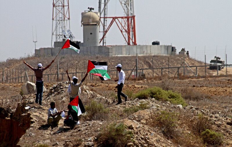 Youths holding Palestinian flags stand in front of Israel's border wall and military jeep