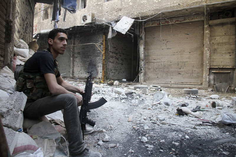 Man holding rifle sits on sandbag in alley littered with debris