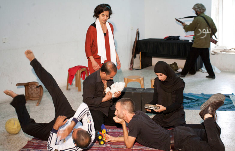 Scene from play shows family sitting together in foreground with Israeli soldier character in background