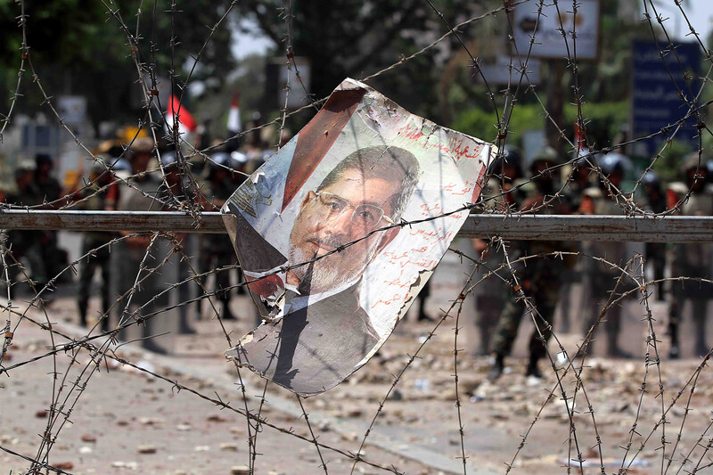 A poster of Mohammed Morsi entangled in barbed wire, with soldiers in background