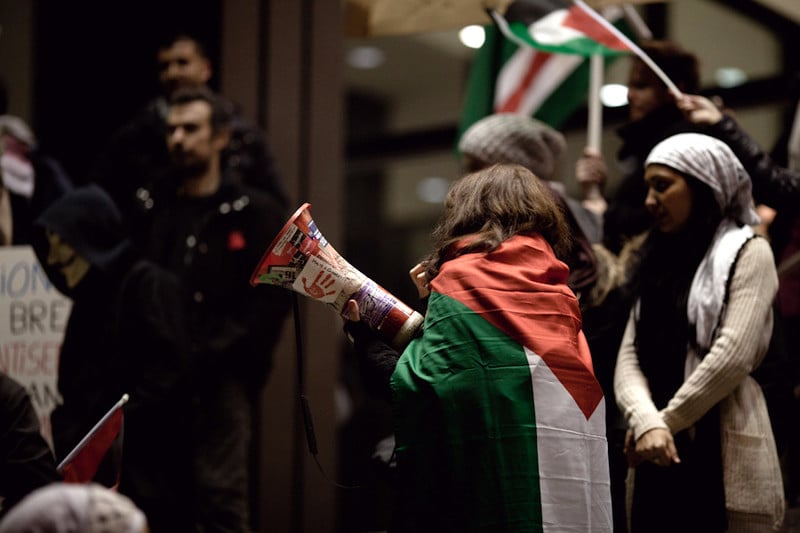 Woman draped in Palestine flag holds a megaphone during nighttime protest