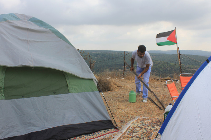 Man picks up bar amid tents with Palestine flag and landscape in background