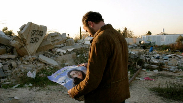 Man looks at the poster he is holding of woman, with rubble in background