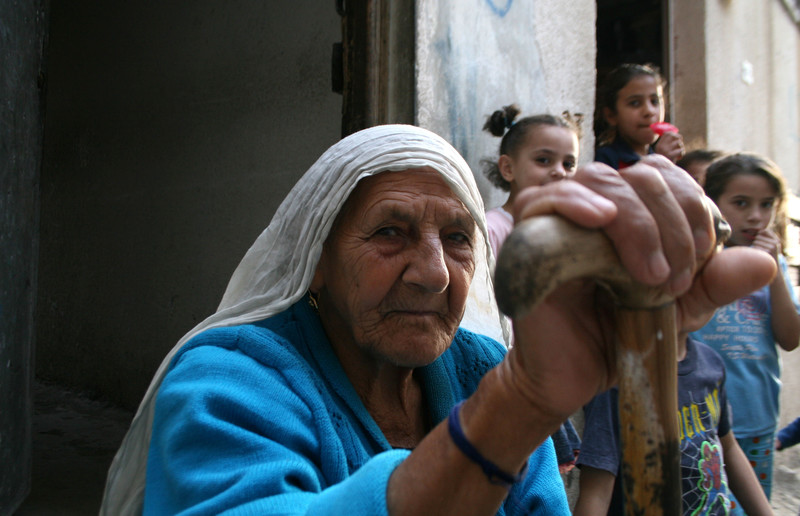 Elderly woman holding cane sits in front of children in alleyway
