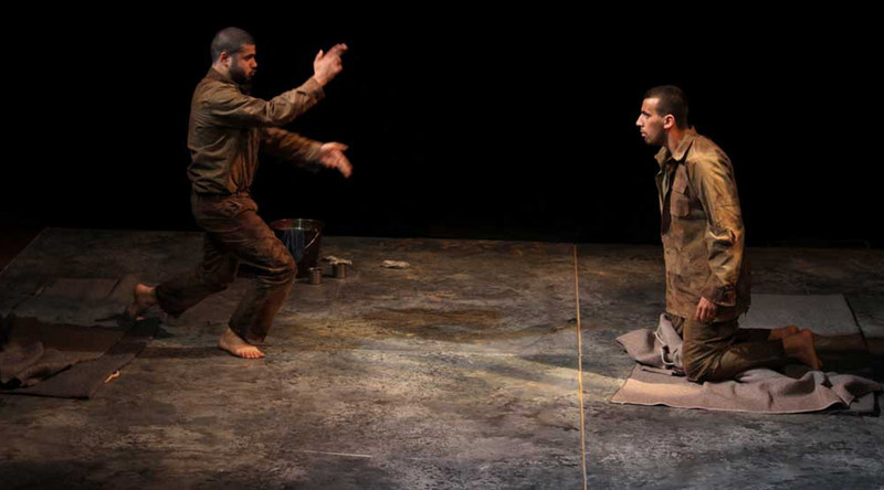 A standing man gestures to a kneeling man on a dark, spare stage