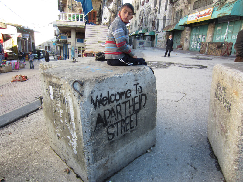 Boy sits on cement block which is spray painted with a stencil reading Welcome to apartheid street