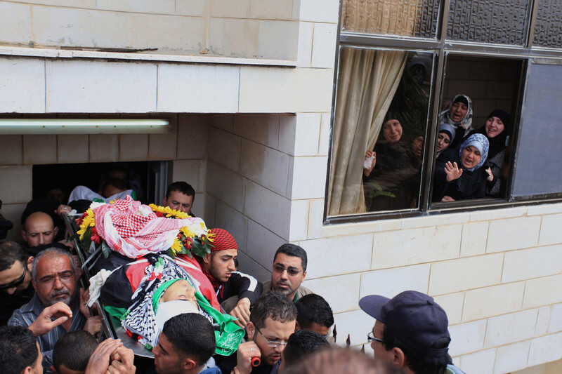 Women looking out of women cry as men carry body of young man out of home in funeral procession