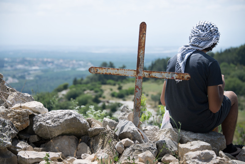 Young man with back to camera sits next to cross on hilltop overlooking valley