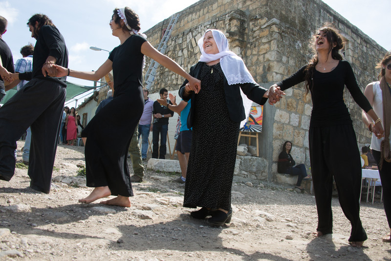 An elderly woman dances with young women in a folk dance line outside old building