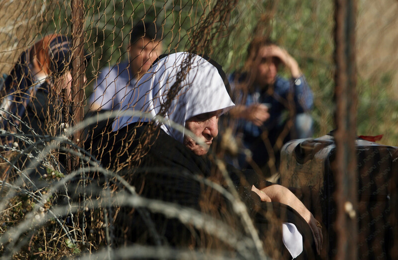 Elderly woman sits next to suitcase near fence and barbed wire