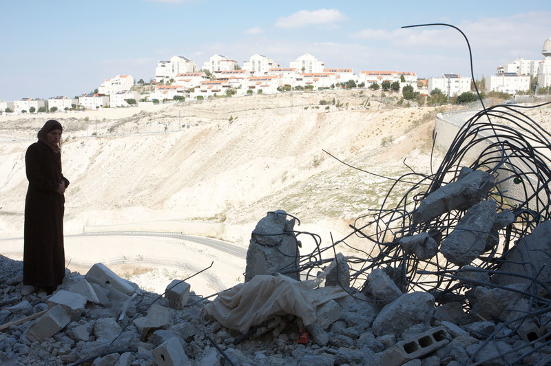 Palestinian woman stands among rubble of destroyed home with Israeli settlement in background