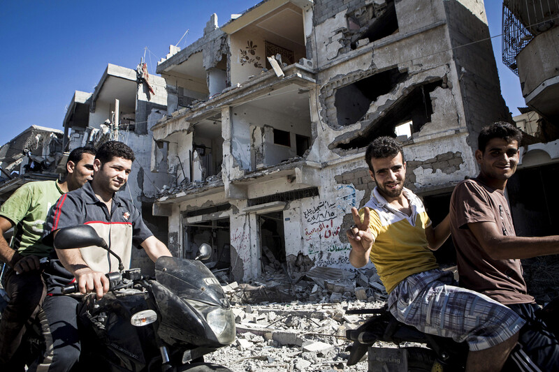 Smiling young men on motorbikes give victory sign in front of shelled building in Gaza City